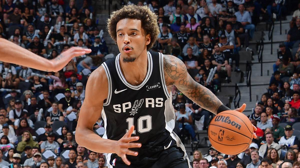 Dwóch koszykarzy podczas meczu, jeden w czarnym stroju z numerem 10 prowadzi piłkę, drugi w czerwonym stroju z numerem 14 próbuje go zatrzymać.