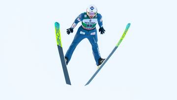 Skoczek narciarski w locie, w niebieskim kombinezonie, z nartami ustawionymi do lądowania.