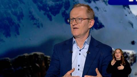 Mężczyzna w okularach i marynarce, m&oacute;wiący z entuzjazmem, na tle zaśnieżonych g&oacute;r.