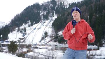 Mężczyzna w czerwonej bluzie i niebieskiej czapce z daszkiem, trzymający mikrofon, stoi na tle ośnieżonych skoczni narciarskich i zimowego krajobrazu.