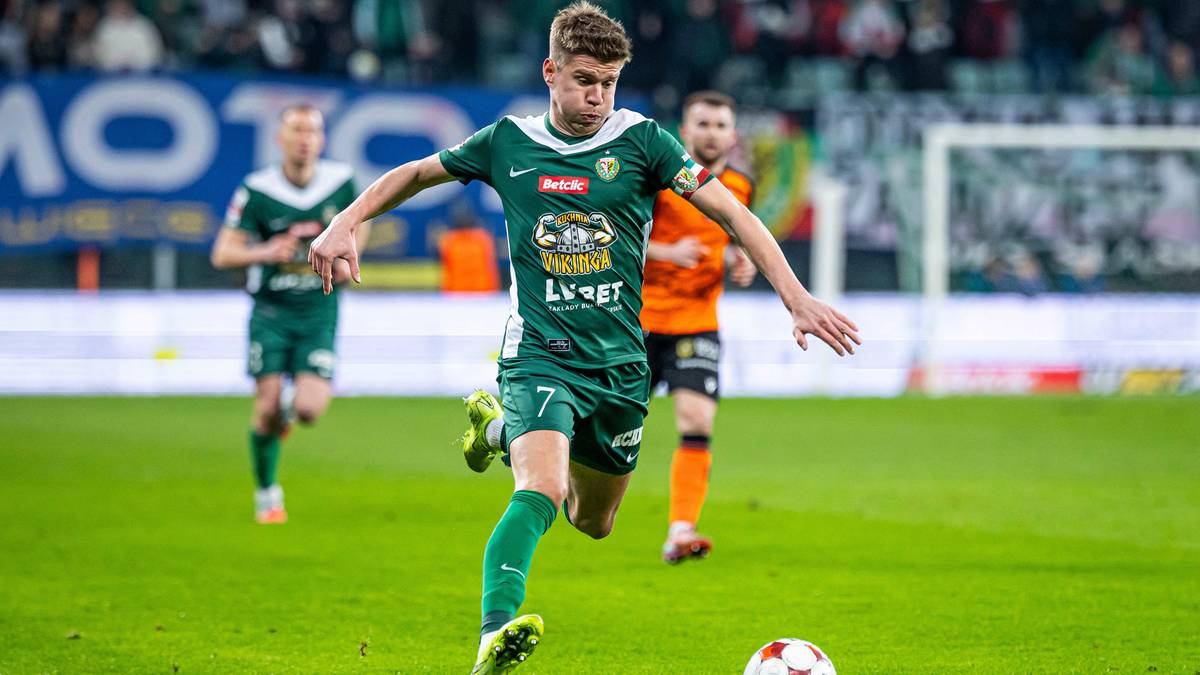 Piłkarz w zielonej koszulce prowadzi piłkę na boisku piłkarskim.