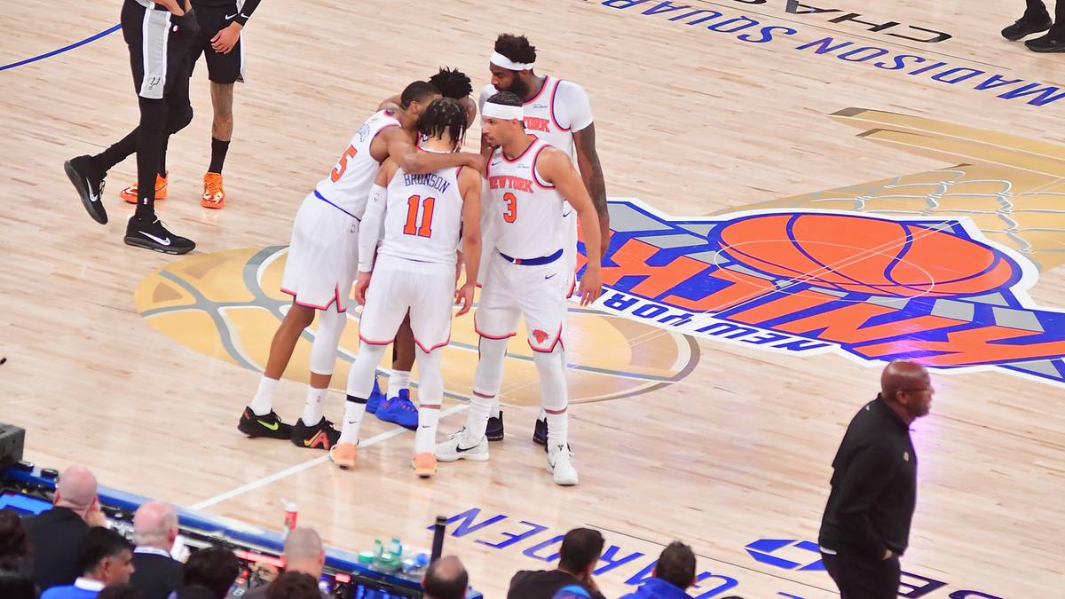 Grupa koszykarzy drużyny New York Knicks w białych strojach z pomarańczowymi detalami, przytulających się na boisku. Na podłodze widoczne jest logo Madison Square Garden.
