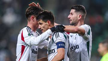 Trzech piłkarzy Legii Warszawa w białych strojach świętuje bramkę, przytulając się do siebie na boisku.