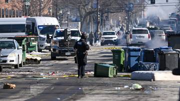 Policjant stoi na ulicy pełnej śmieci i porozrzucanych przedmiot&oacute;w. W tle widać samochody i budynki.