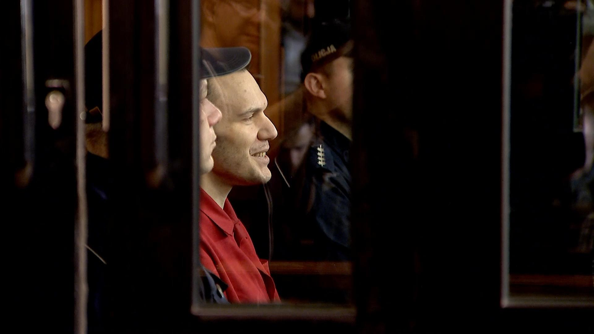 Stefan Wilmont siedzi za kratami sali sądowej, ubrany w czerwoną koszulę, z widocznym uśmiechem.