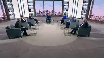 Siedem os&oacute;b siedzi na kanapach w studiu telewizyjnym, dyskutując. W tle widać okna z widokiem na panoramę miasta.