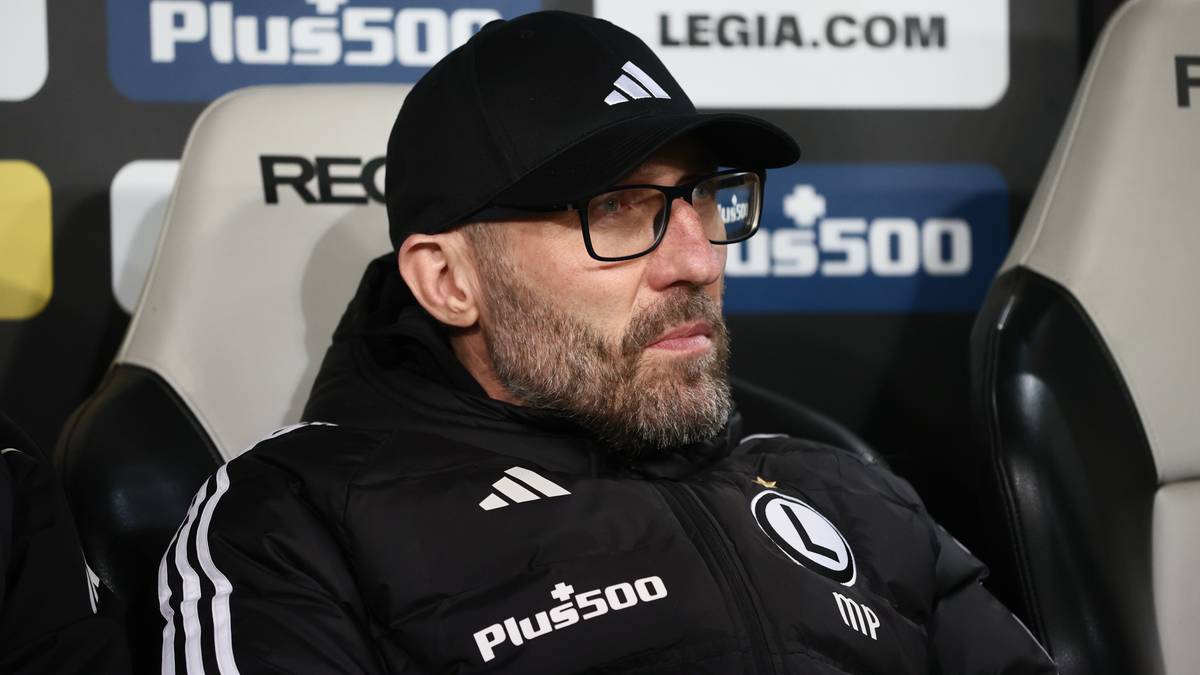 Trener Legii Warszawa w czapce z daszkiem i okularach siedzi na ławce rezerwowych.