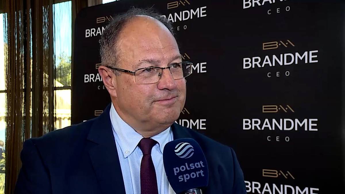 Marian Kmita w okularach i granatowym garniturze z bordowym krawatem, trzyma mikrofon Polsat Sport przed ścianką z logo BrandMe CEO.