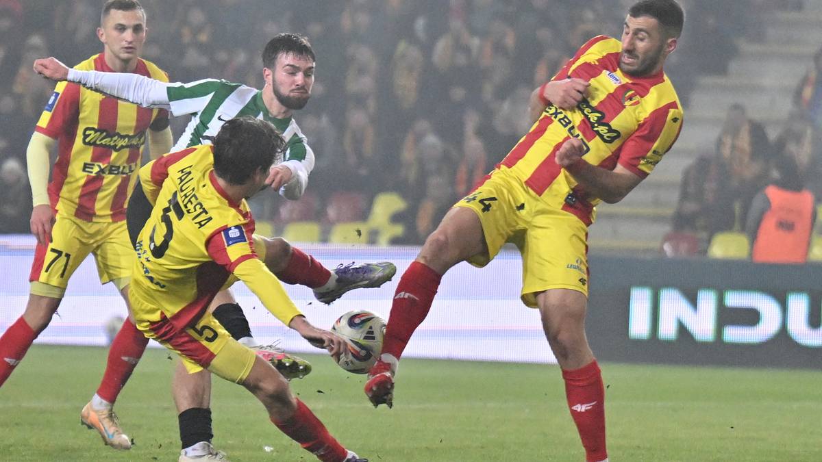 Piłkarze w ż&oacute;łto-czerwonych strojach walczą o piłkę z zawodnikiem w zielono-białym stroju.