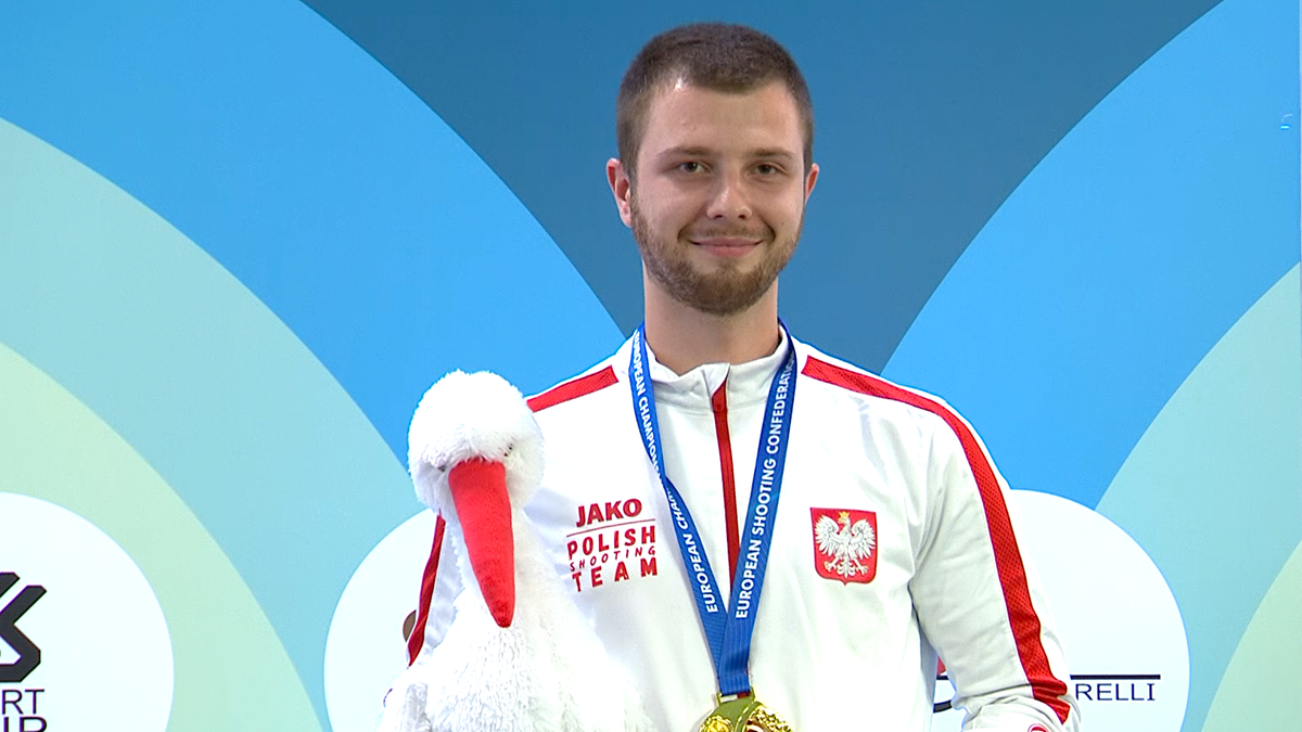 Mężczyzna w białej koszulce z czerwonymi rękawami i napisem "Jako Polish Shooting Team", z medalem na szyi, trzymający pluszowego bociana.