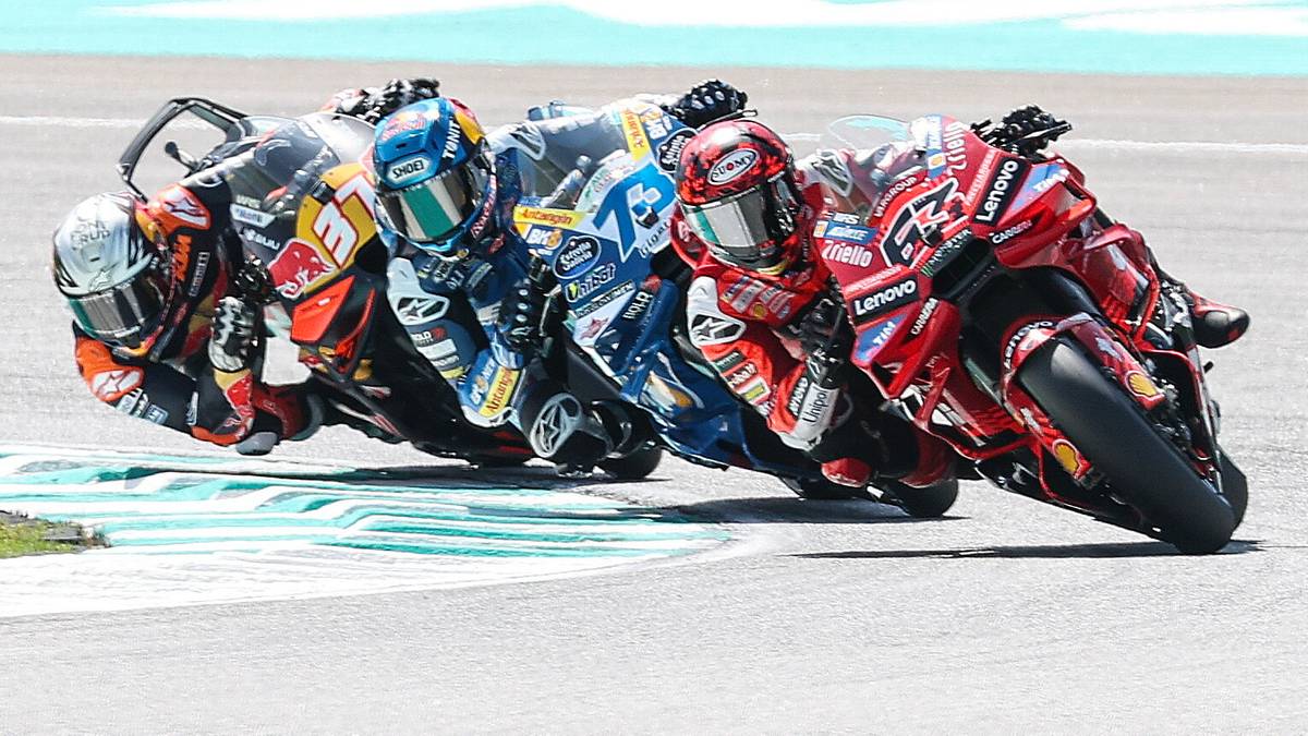 Grupa motocyklist&oacute;w w kaskach i kombinezonach ściga się na torze wyścigowym, pochyleni w zakręcie.