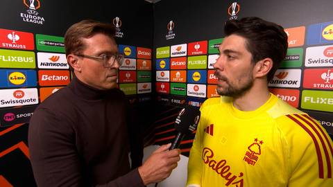 Dw&oacute;ch mężczyzn rozmawia ze sobą. Jeden z nich, ubrany w ż&oacute;łtą koszulkę z logo drużyny piłkarskiej, jest przepytywany przez drugiego, kt&oacute;ry trzyma mikrofon. W tle widać ściany z logo sponsor&oacute;w.