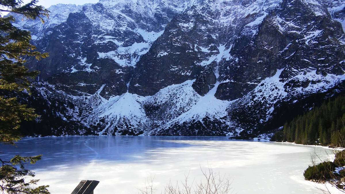 Zamarznięte Morskie Oko w otoczeniu ośnieżonych g&oacute;r, z widocznym drewnianym płotem na pierwszym planie.