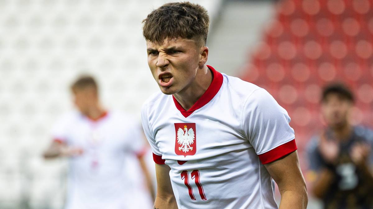 Młody piłkarz w białej koszulce z godłem Polski i numerem 11 na piersi, z zacisniętymi ustami i wyrazem determinacji na twarzy, podczas meczu.