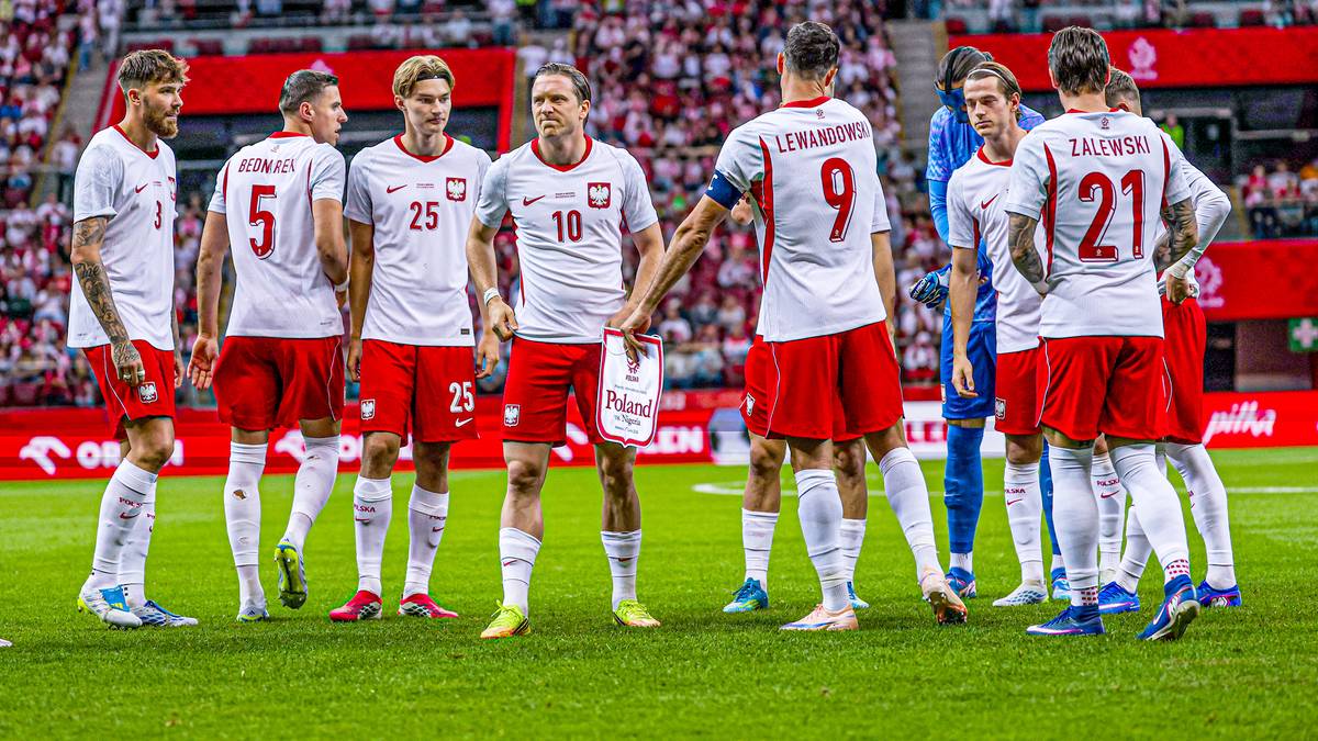 Piłkarze reprezentacji Polski w białych koszulkach i czerwonych spodenkach stoją na zielonej murawie stadionu.