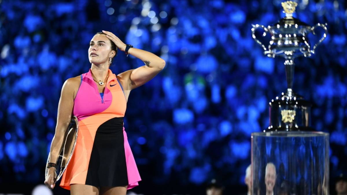 Tenisistka Aryna Sabalenka stoi na korcie, trzymając rakietę, z ręką na czole, patrząc na trofeum.