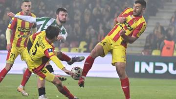 Piłkarze w ż&oacute;łto-czerwonych strojach walczą o piłkę z zawodnikiem w zielono-białym stroju.