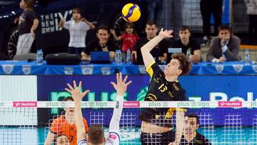 Siatkarz w ż&oacute;łto-czarnej koszulce z numerem 13 atakuje piłkę nad siatką, podczas gdy inny zawodnik w białej koszulce pr&oacute;buje go zablokować.