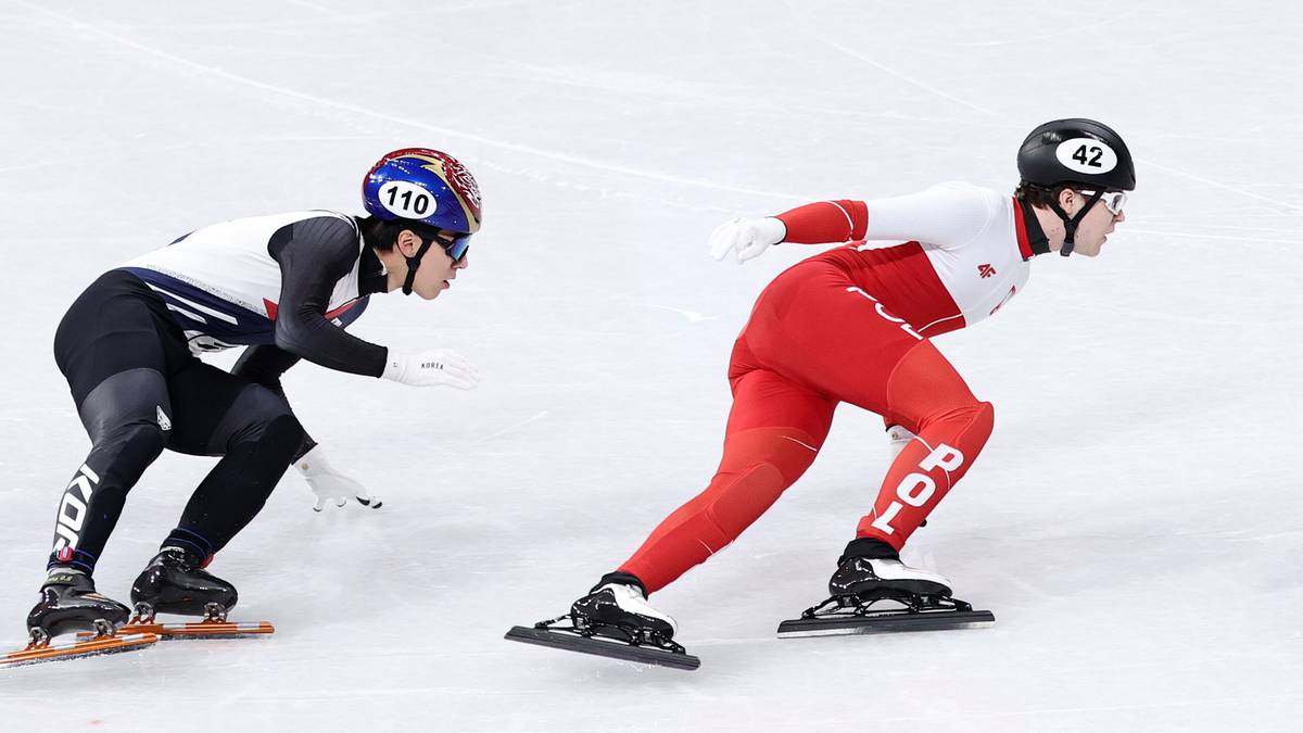 Trzech zawodnik&oacute;w short tracka w kaskach i strojach sportowych ściga się na lodowisku.