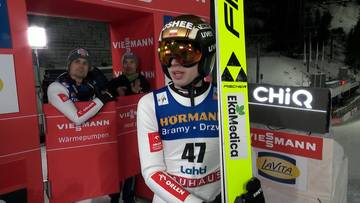 Skoczek narciarski w stroju sportowym i kasku, trzymający nartę, stoi przed zamazanym tłem z innymi osobami i logotypami.