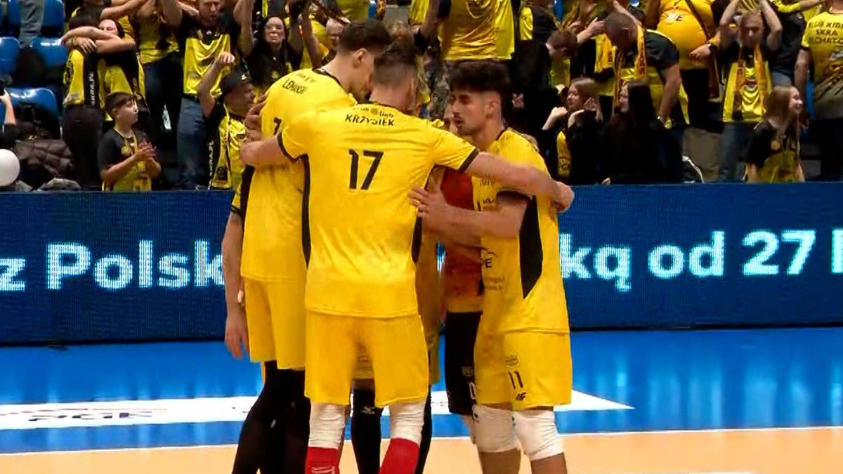 Sześciu siatkarzy w żółtych strojach sportowych stoi w kręgu, obejmując się nawzajem po meczu. Na koszulkach widoczne są numery. W tle widać kibiców na trybunach.