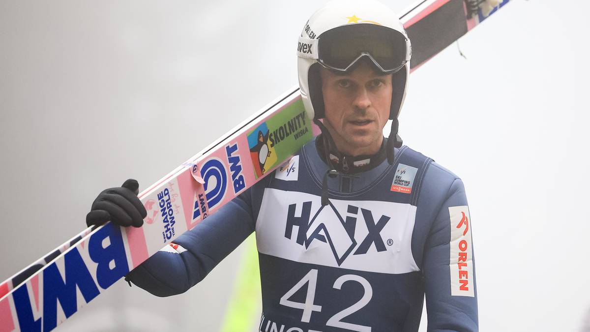 Skoczek narciarski w kasku i goglach, trzymający narty na ramieniu. Ma na sobie niebiesko-biały kombinezon z numerem 42.