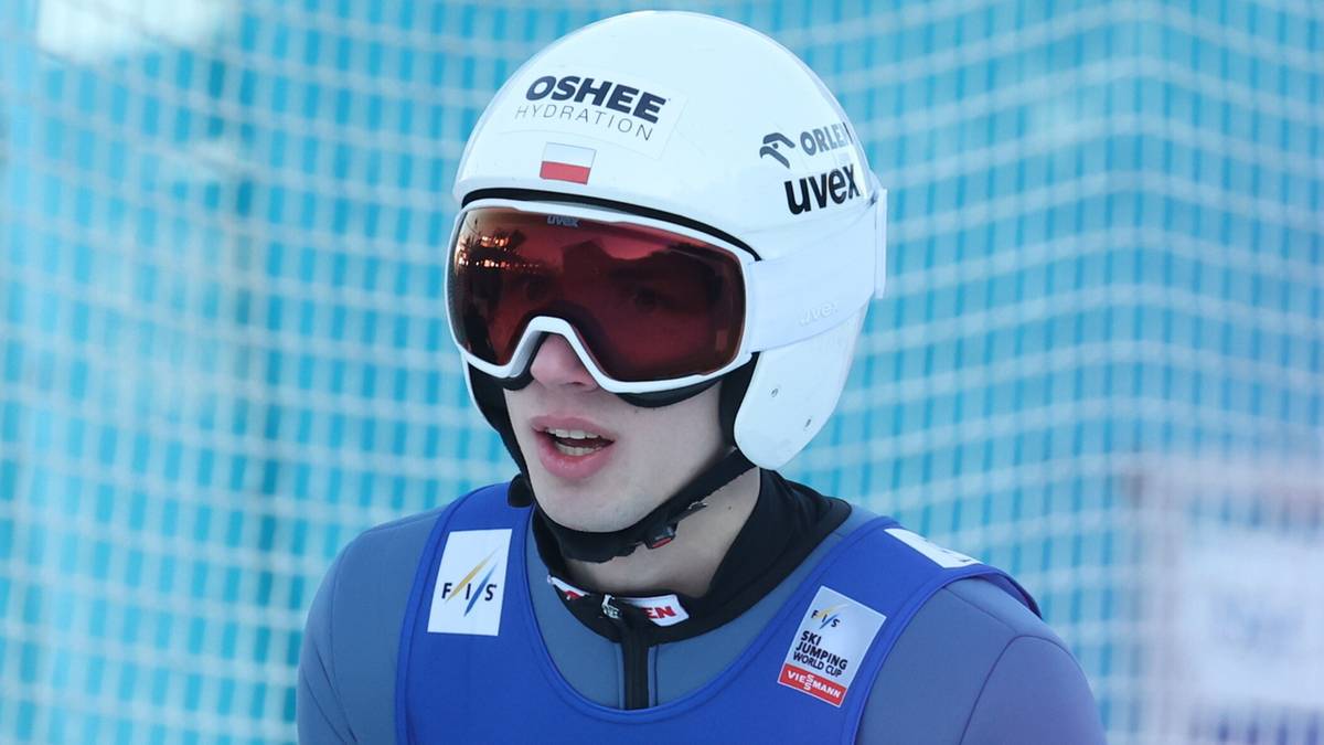 Skoczek narciarski w biało-niebieskim kombinezonie z numerem 19 na piersi i w goglach narciarskich.