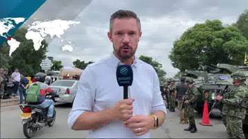 Mężczyzna w białej koszuli trzyma mikrofon i m&oacute;wi do kamery. Za nim widać wojskowe pojazdy i żołnierzy. W tle znajduje się też mapa świata z zaznaczonym obszarem.