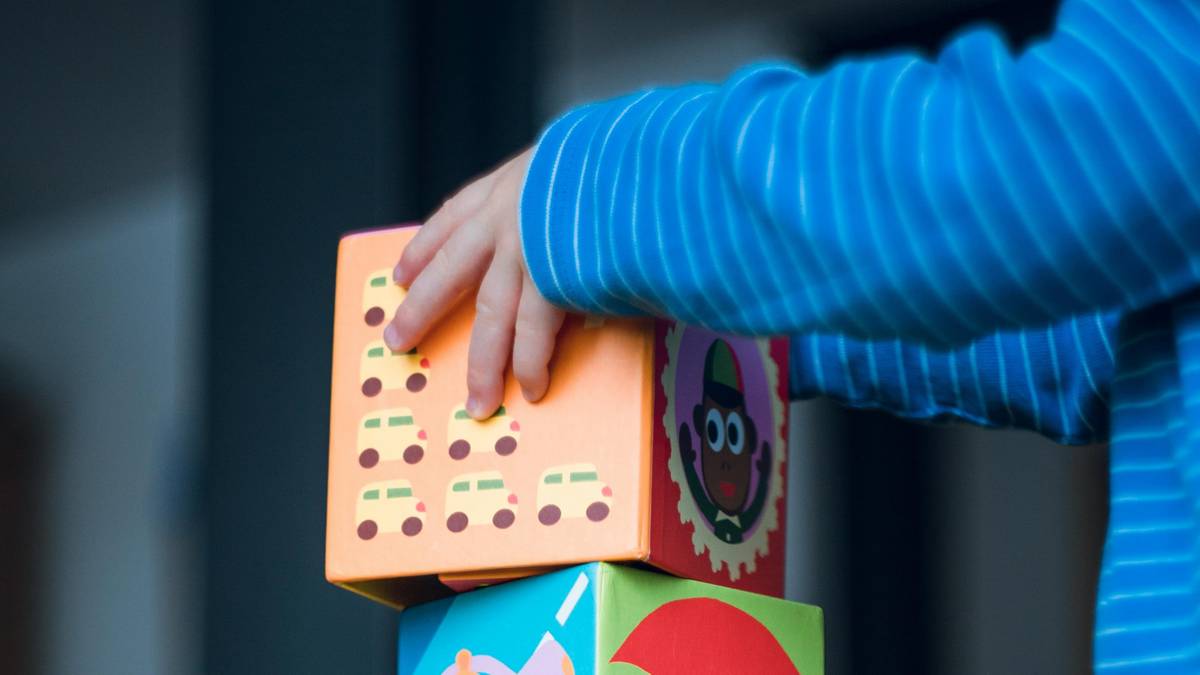 Dłoń dziecka dotyka pomarańczowego klocka z wizerunkami samochodzików, obok znajdują się inne kolorowe klocki.