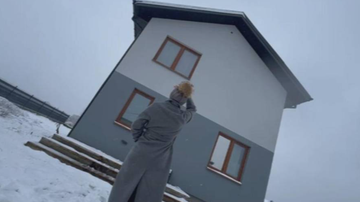 Kobieta w szarym płaszczu stoi przed nowoczesnym domem o szarej i białej elewacji, pokrytym śniegiem. Widoczne są drewniane okna i pochmurne niebo.