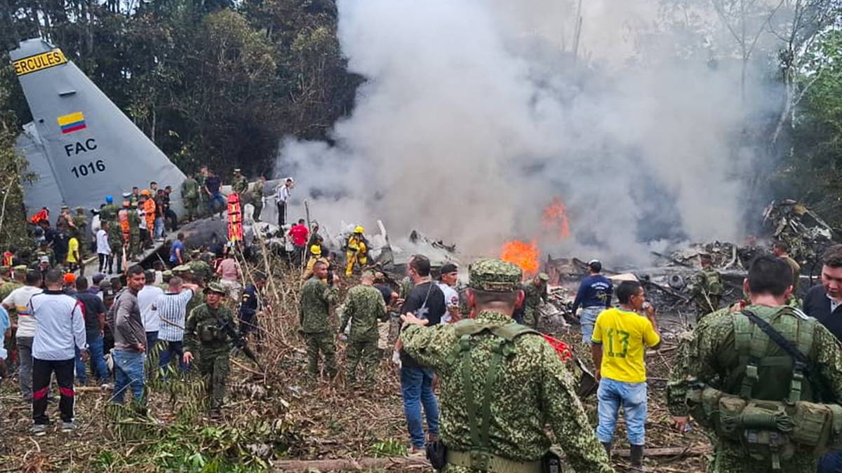 Widok na miejsce katastrofy wojskowego samolotu Hercules C-130 z płonącymi szczątkami i dymem, otoczone przez osoby w mundurach i cywilne.