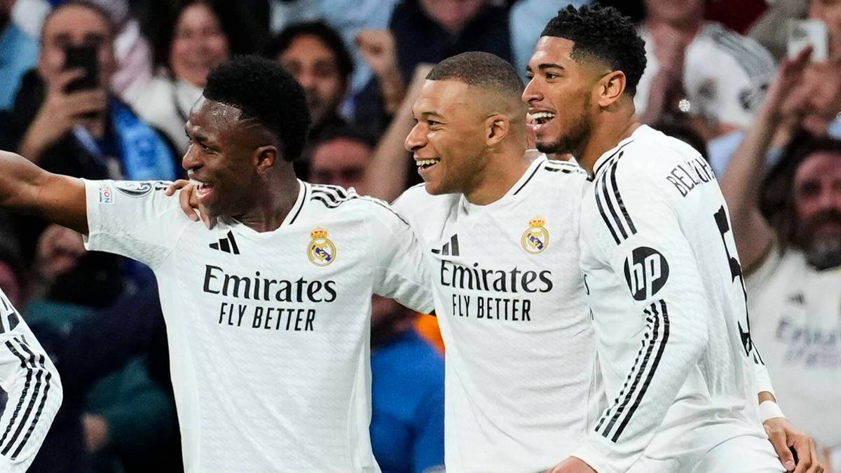 Trzech piłkarzy Realu Madryt w białych strojach, Vinícius Júnior, Kylian Mbappé i Éder Militão, uśmiecha się i cieszy ze zdobytej bramki, z kibicami w tle.