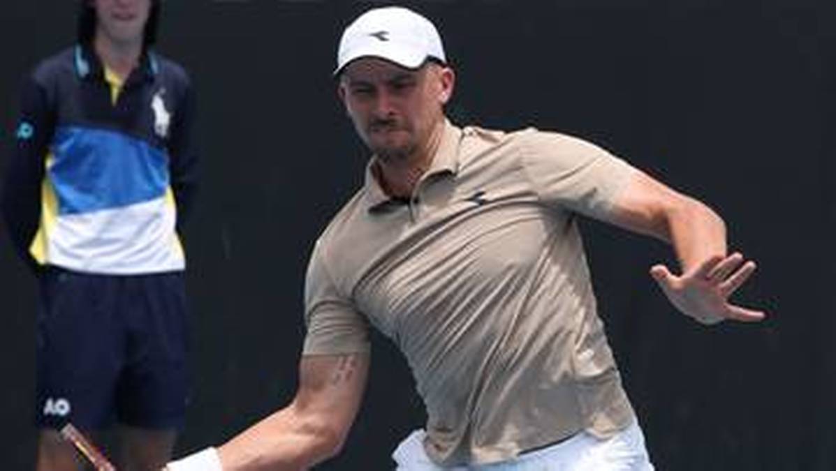 Dwóch mężczyzn w strojach tenisowych na korcie. Na pierwszym planie zawodnik w beżowej koszulce i białej czapce z daszkiem wykonuje zamach rakietą. W tle stoi drugi mężczyzna w granatowej spódnicy i bluzie z kolorowymi pasami.