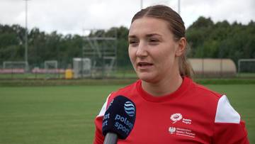 Młoda kobieta w czerwonej koszulce sportowej z białymi rękawami, z mikrofonem przed ustami, mówi do kamery. W tle boisko sportowe.