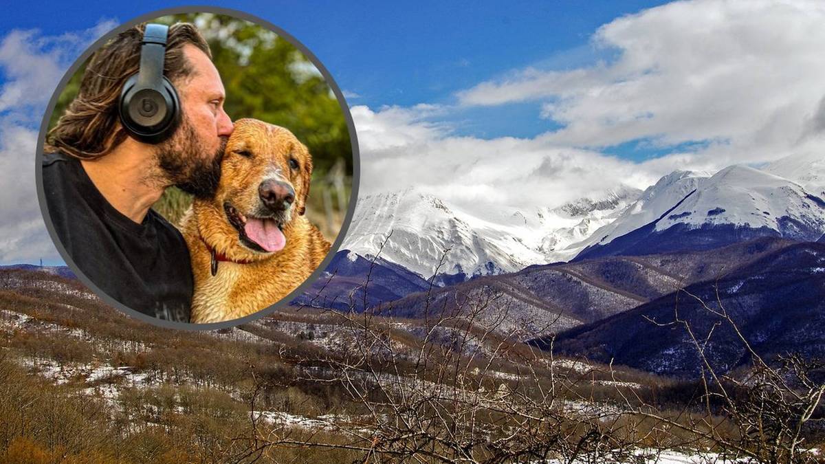 Mężczyzna w słuchawkach całuje psa w policzek na tle górskiego krajobrazu.