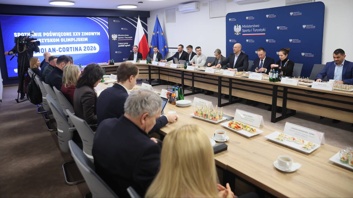 Grupa os&oacute;b siedzi przy długim stole konferencyjnym, przed nimi leżą poczęstunki. Na ekranie za nimi wyświetlany jest napis "Polska na Igrzyskach Olimpijskich Pekin 2022".