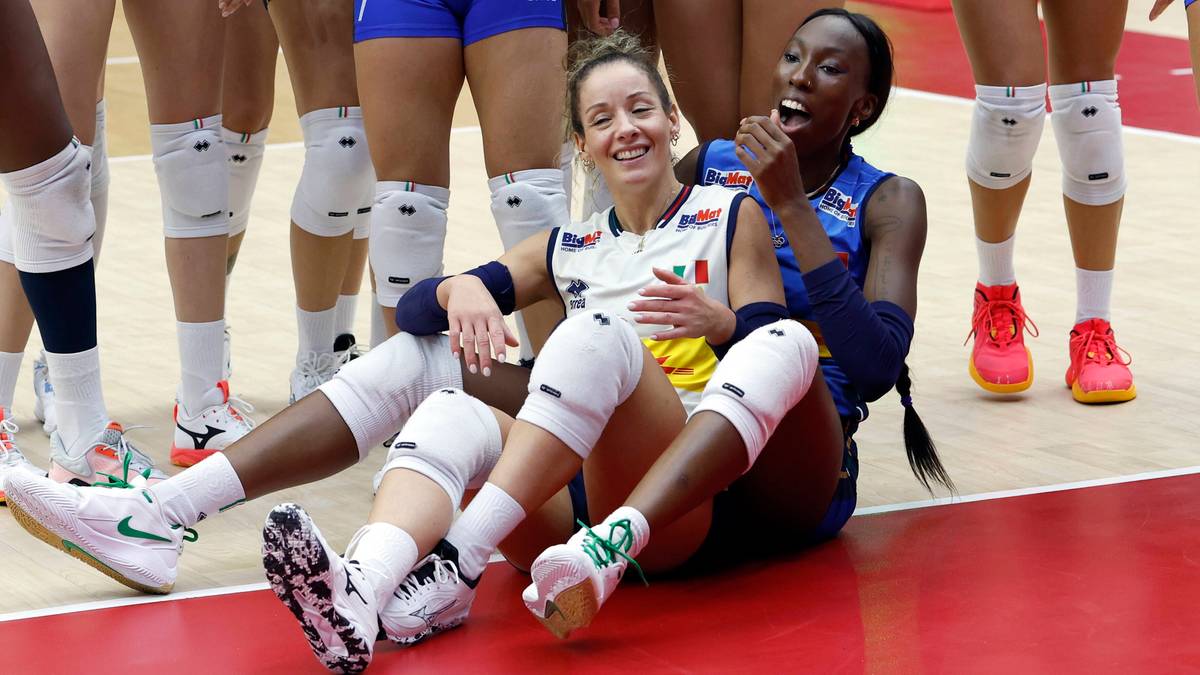 Dwie siatkarki siedzą na podłodze w strojach sportowych, jedna z nich uśmiecha się szeroko, druga jest za nią.