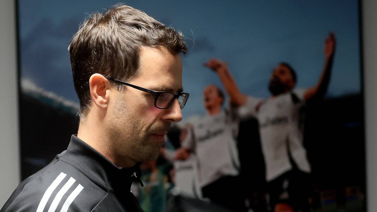 Mężczyzna w okularach i czarnej bluzie z białymi paskami z bliska po lewej stronie, z rozmazanym tłem przedstawiającym piłkarzy świętujących zwycięstwo.