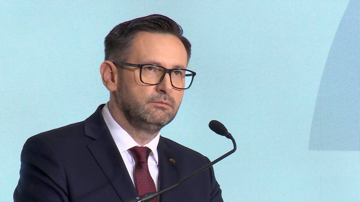 Portret mężczyzny w okularach i garniturze, m&oacute;wiącego do mikrofonu.