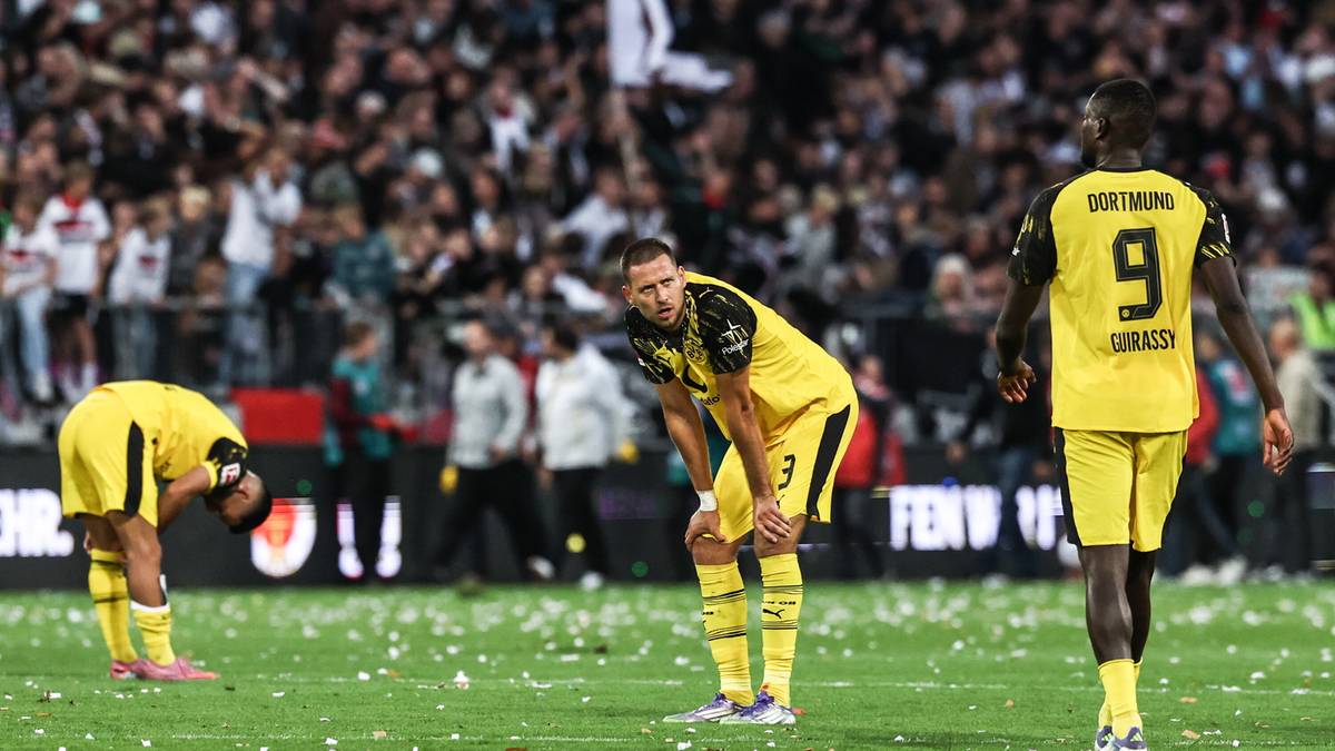 Trzech piłkarzy w żółtych strojach Borussii Dortmund na boisku piłkarskim, dwaj z nich pochyleni z rezygnacją, jeden z nich z numerem 9 na plecach stoi z opuszczoną głową. W tle trybuny z kibicami.