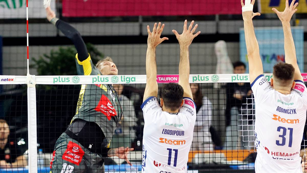 Siatkarz w ż&oacute;łto-czarnej koszulce wykonuje atak nad siatką, blokowany przez dw&oacute;ch zawodnik&oacute;w w białych koszulkach.