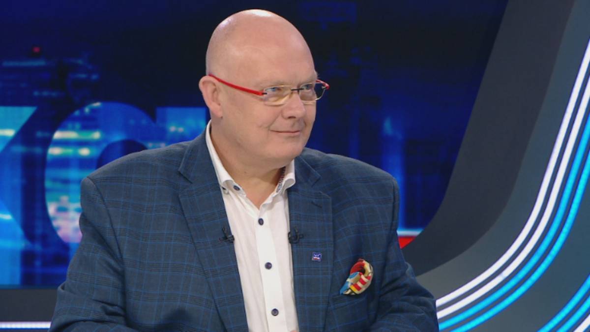 Mężczyzna w średnim wieku, łysiejący, w okularach i marynarce, siedzi w studiu telewizyjnym.