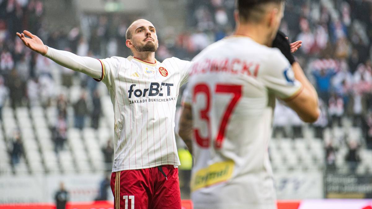 Piłkarz w białej koszulce z czerwonymi elementami świętuje zdobycie bramki z uniesionymi rękami.