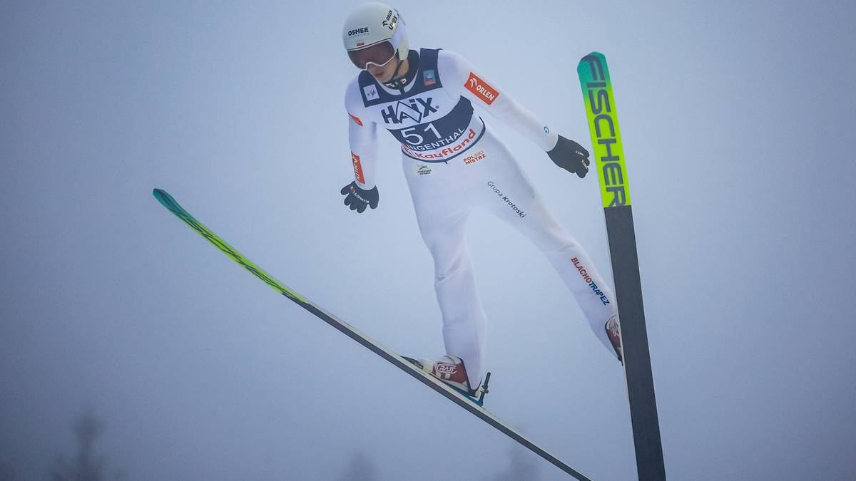 Skoczek narciarski w białym kombinezonie i kasku w locie na tle mglistego krajobrazu.