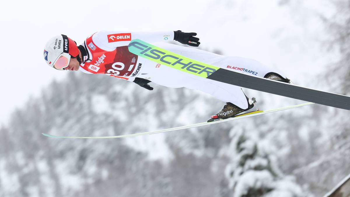 Skoczek narciarski w locie, w białym kombinezonie i z nartami Fischer, na tle zaśnieżonych drzew.