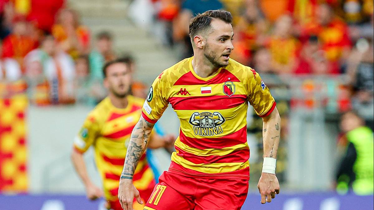 Piłkarz Jagiellonii Białystok w żółto-czerwonej koszulce z herbem klubu, biegnący po boisku, z widownią w tle.