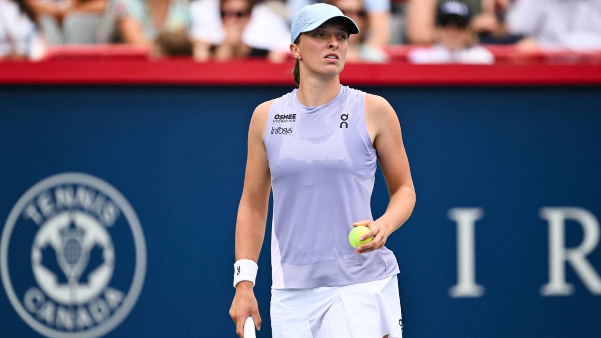 Iga Świątek, polska tenisistka, w pastelowej koszulce bez rękawów i białej spódniczce, z czapką z daszkiem na głowie, trzyma zieloną piłkę tenisową w dłoni, stojąc na korcie tenisowym z niebieskim tłem z logo.