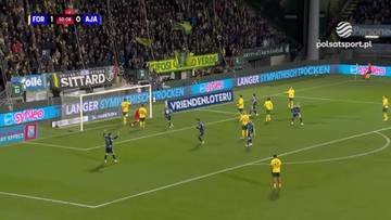 Piłkarze w żółtych i niebieskich strojach rywalizują na boisku piłkarskim podczas meczu Fortuna Sittard - Ajax Amsterdam. W tle widać trybuny wypełnione kibicami.