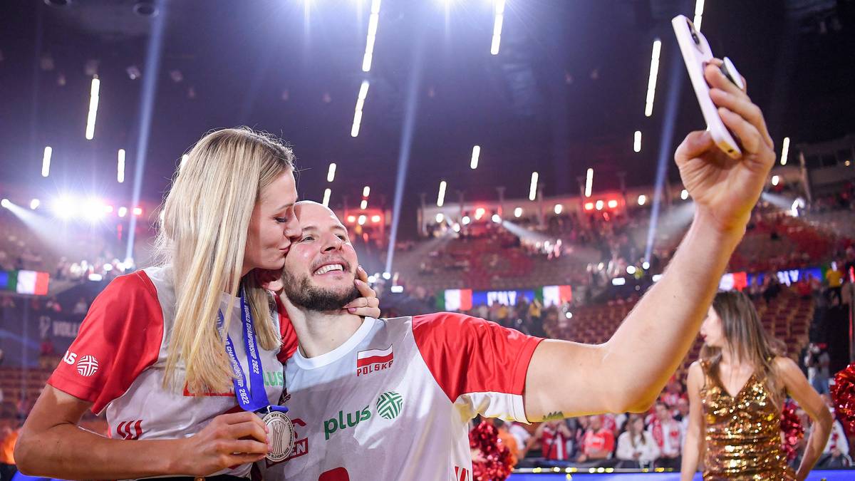 Anna Kurek całuje swojego męża Jakuba w policzek, podczas gdy on robi sobie zdjęcie telefonem komórkowym. Oboje są ubrani w biało-czerwone stroje sportowe.