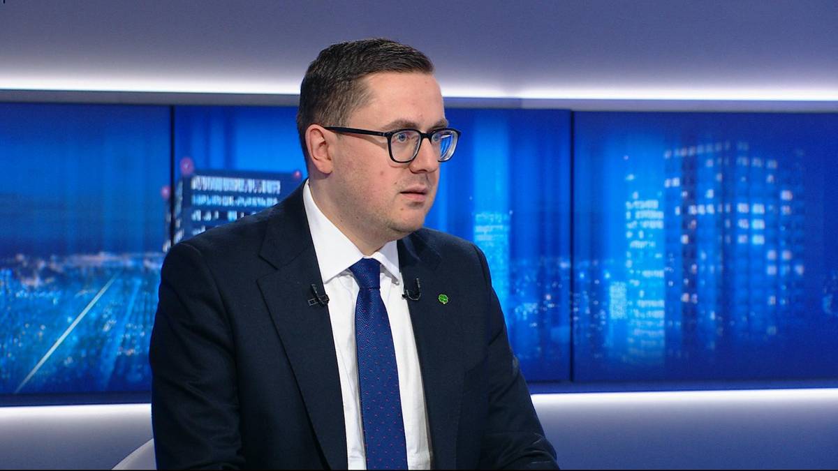 Mężczyzna w okularach i garniturze siedzi przed niebieskim tłem.